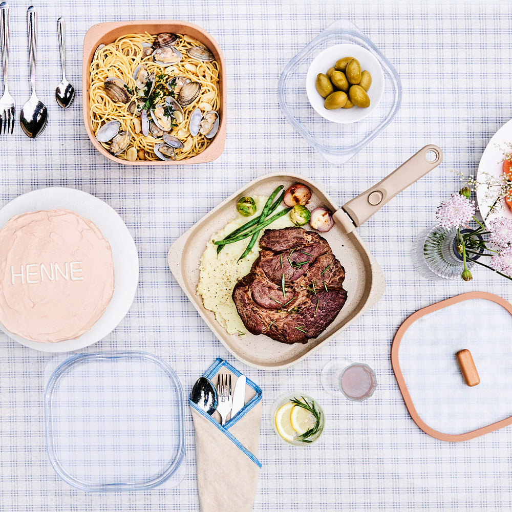 Top view of a table set with grilled meat, pasta, and a beige skillet with a detachable handle.