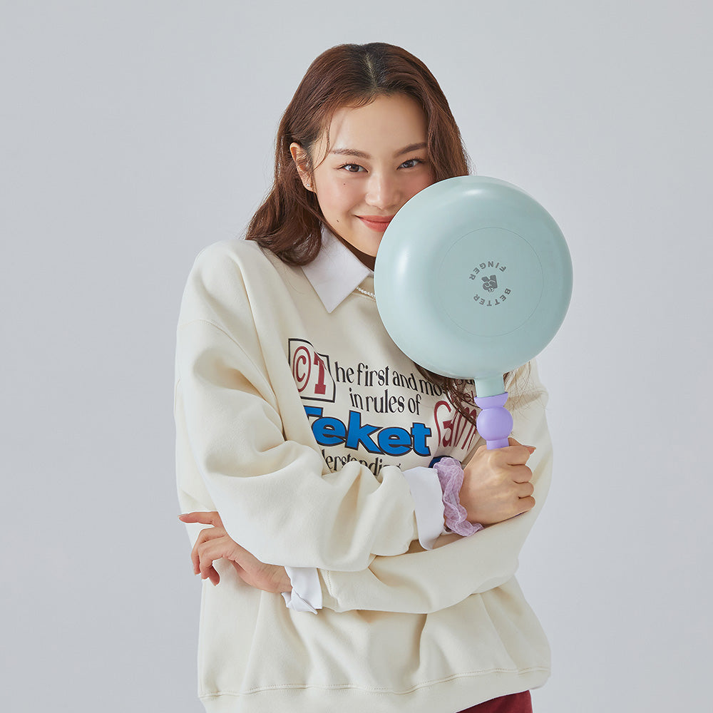 A woman in a cream sweatshirt holding a light green frying pan with a purple handle, smiling and posing against a light gray background