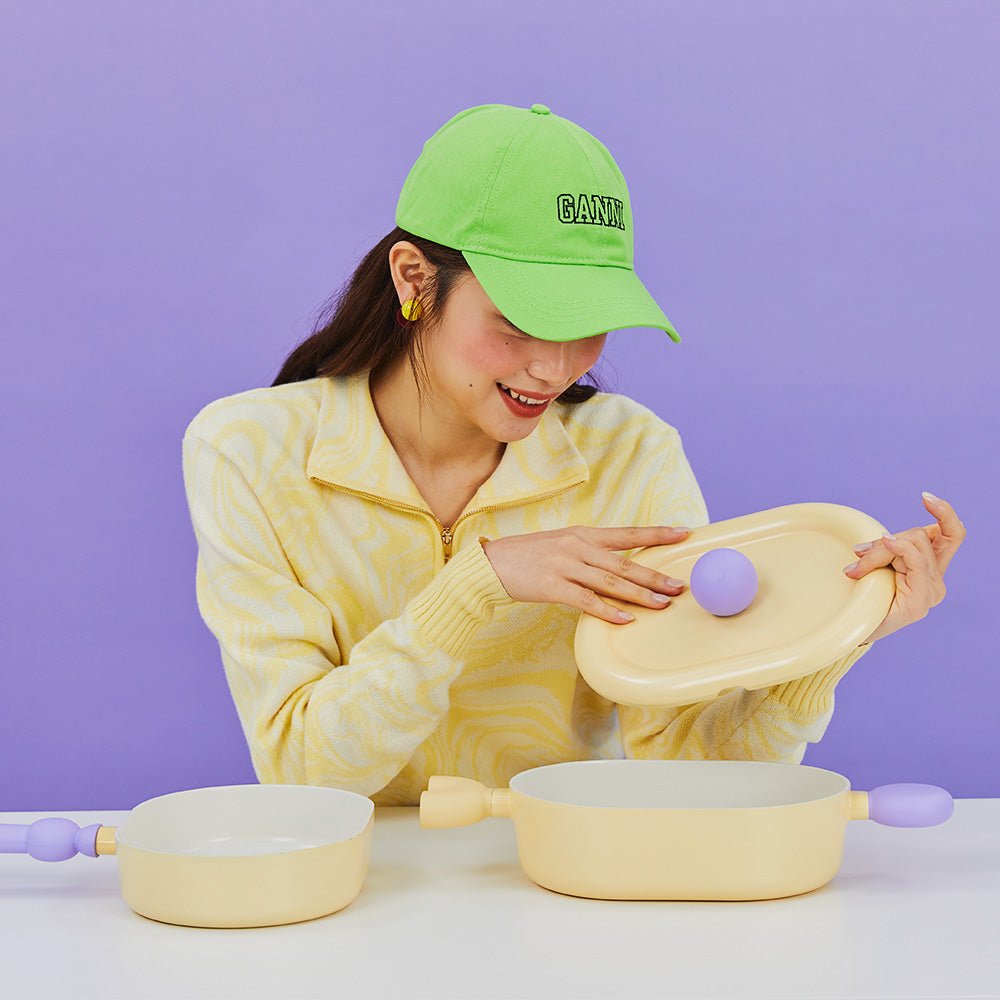 A woman in a yellow sweater and green cap, smiling as she holds the lid of a yellow oval pot with purple handles, sitting at a table against a purple background